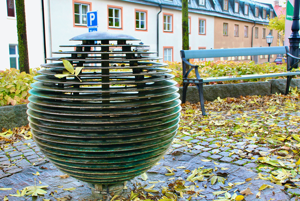 Foto på en fontän på Kaplan. Fontänen ser ut som en boll gjord i metallskivor. Runt fontänen syns massor av höstlöv. I bakgrunden bakom fontänen syns en parkbänk och en plantering med sockblommor och träd.