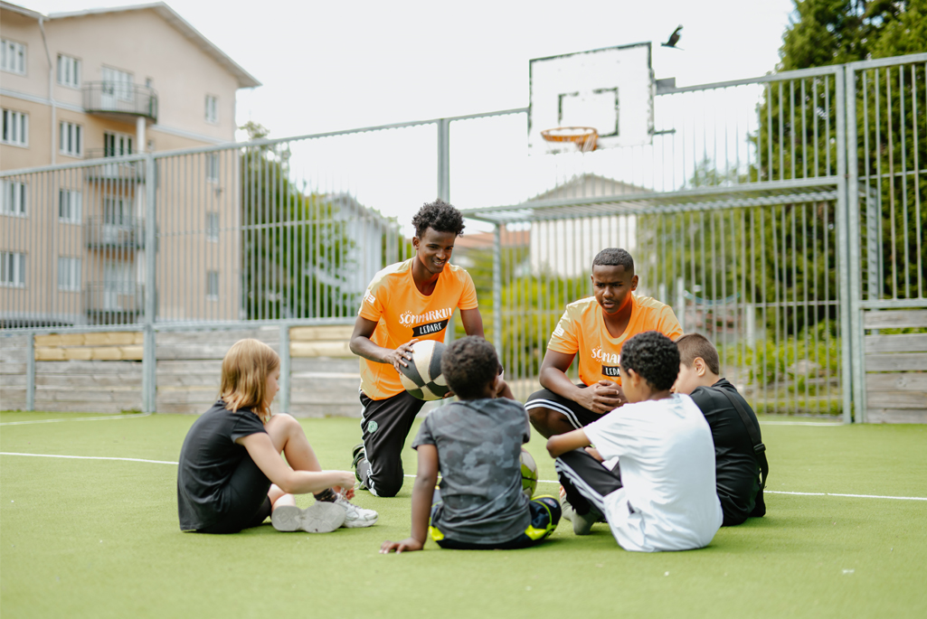 Ungdomar och barn som sitter i en ring på en basketplan med en basketboll och två ledare. Foto: Sommarkul